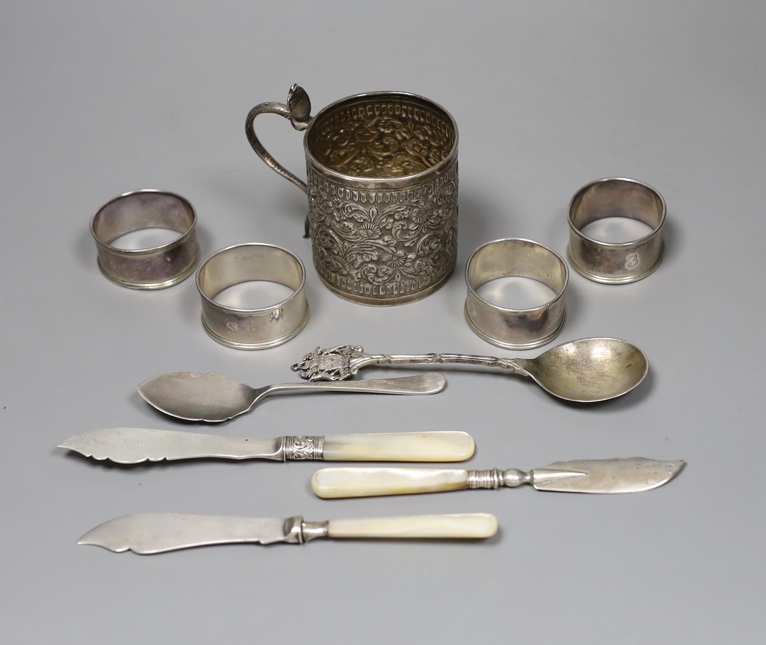 A 19th century Indian white metal christening can with cobra handle, a set of four silver napkin rings, two spoons and three mother of pearl handled butter knives.